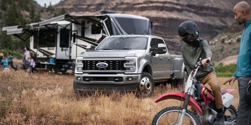 
Silver Ford Super Duty dually towing an RV at a campsite with a child on a dirt bike